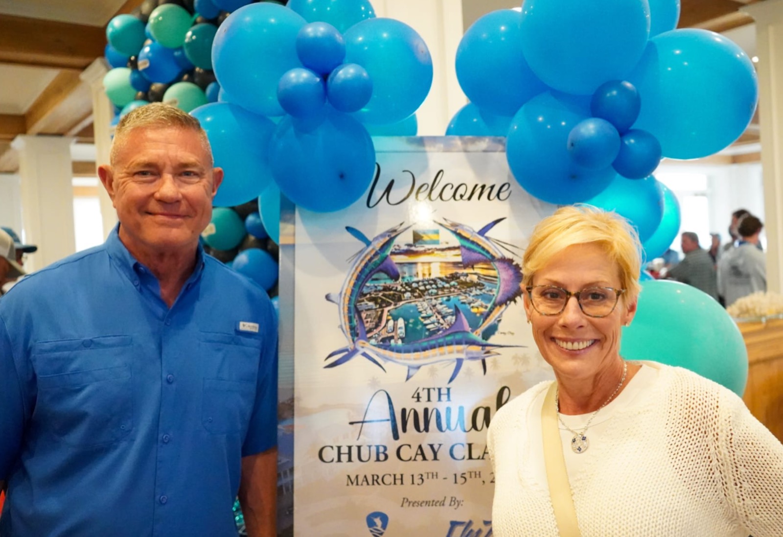 Sam and Jennifer Galloway smiling in front of a Chub Cay Classic welcome sign and blue balloons in the Clubhouse